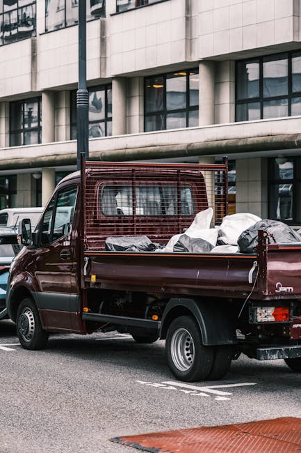 A maroon flatbed pickup truck parked on an urban street in front of a modern office building with large glass windows and a concrete facade. The truck's open cargo bed is filled with various waste ite